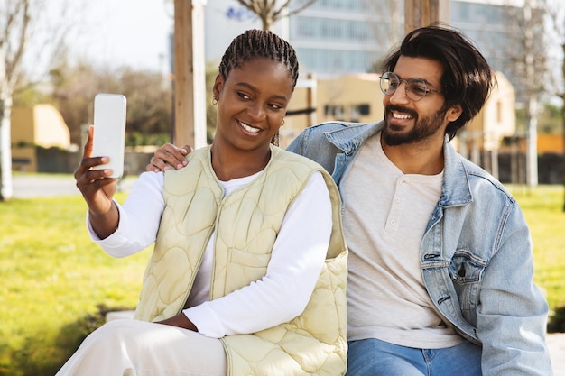 Los estudiantes árabes negros jóvenes alegres estudian juntos con un teléfono inteligente y se reúnen al aire libre para hacer selfie