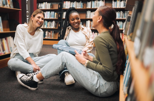 Estudiantes de educación o amigas en el piso de la biblioteca para el aprendizaje de conversaciones cómicas o el estudio de libros Diversidad feliz o niña con una sonrisa para la investigación universitaria de apoyo universitario o proyecto escolar