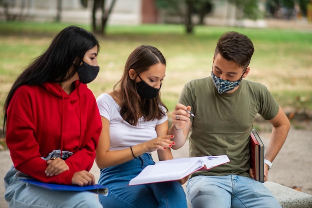 Estudiantes estudiando juntos sentados en un banco al aire libre y usando máscaras covid