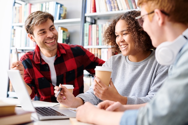 Estudiantes interculturales felices en ropa casual discutiendo videos en línea mientras toman café en la biblioteca