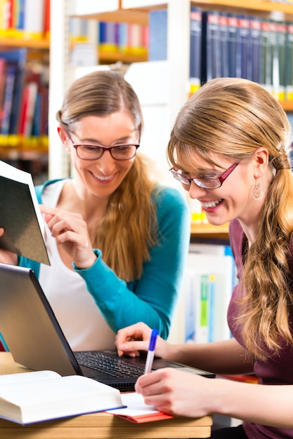 Estudiantes: mujeres jóvenes en biblioteca con ordenador portátil y libros de aprendizaje en grupo