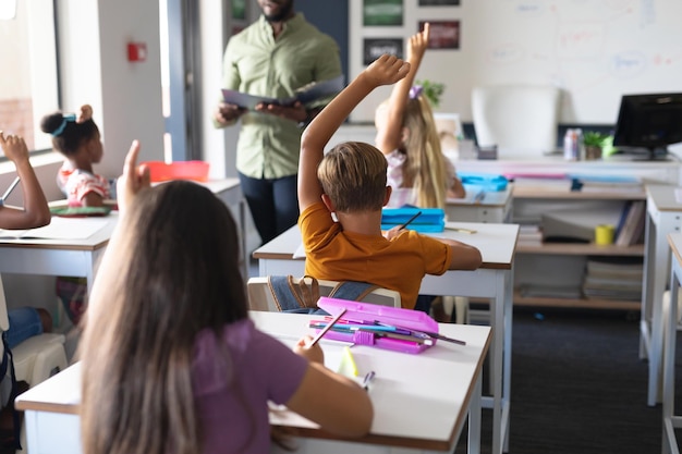 Estudiantes multirraciales levantando la mano mientras el joven maestro afroamericano enseña en el aula. sin alteración, educación, aprendizaje, infancia, enseñanza, ocupación y concepto escolar.