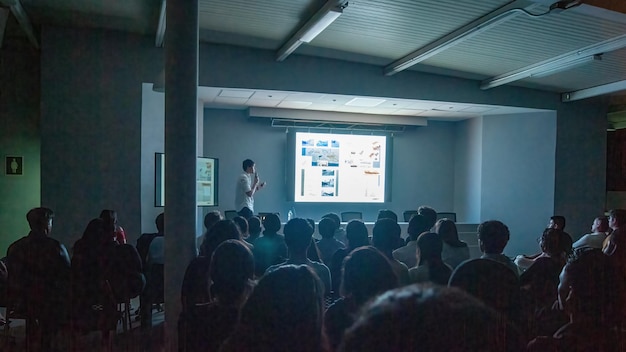 Estudiantes prestando atención a una conferencia impartida por una mujer que proyecta sobre libros y educación arquitectónica