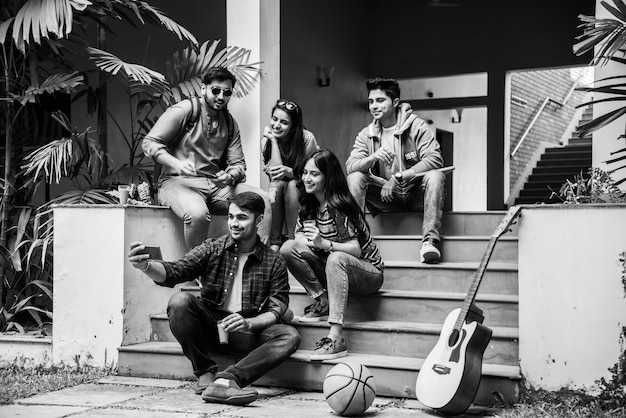 Foto estudiantes universitarios indios asiáticos tomando fotos selfie con smartphone en campus mientras están sentados en las escaleras o el césped, divirtiéndose