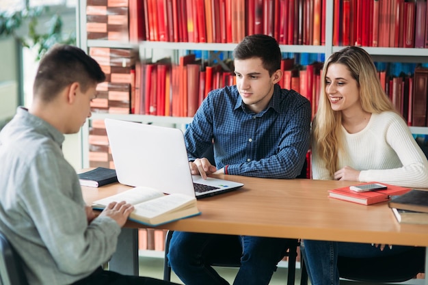 Estudiantes universitarios sentados juntos en la mesa con libros y una computadora portátil Jóvenes felices haciendo estudio grupal en la biblioteca