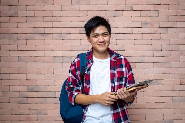 Los estudiantes varones asiáticos usan camisas a cuadros. De pie junto a una pared de ladrillos, cargando una mochila, cargando libros, útiles escolares, preparándose para estudiar, sonriendo.