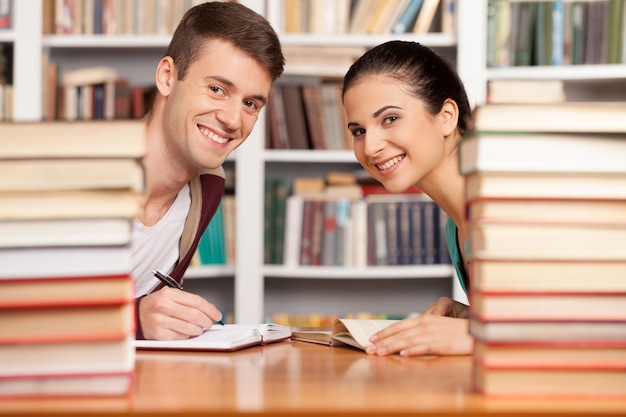 Estudiar juntos. Alegre joven y mujer sentada en el mostrador de la biblioteca y mirando por las pilas de libros