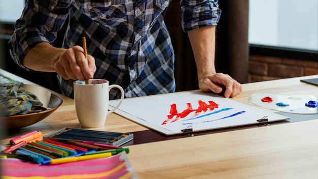 Estudio de artista Pintura de hombre Sumergir pincel en taza Trazos de pincel rojo azul sobre papel Técnica de talento de imaginación
