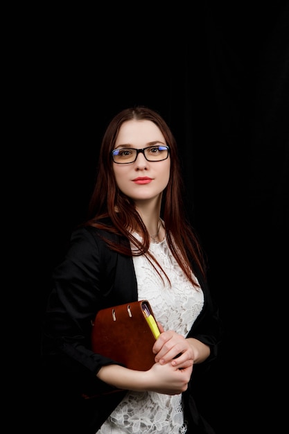 Estudio de cerca el retrato de una niña de apariencia eslava usa chaqueta negra y gafas sobre un fondo negro con un cuaderno marrón en sus manos.