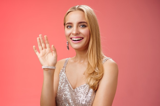 Estudio de cintura para arriba tiro amigable atractiva elegante tierna mujer rubia saludando con la mano levantada saludo acogedor amigo sonriendo encantada presentarse hola gesto, fondo rojo