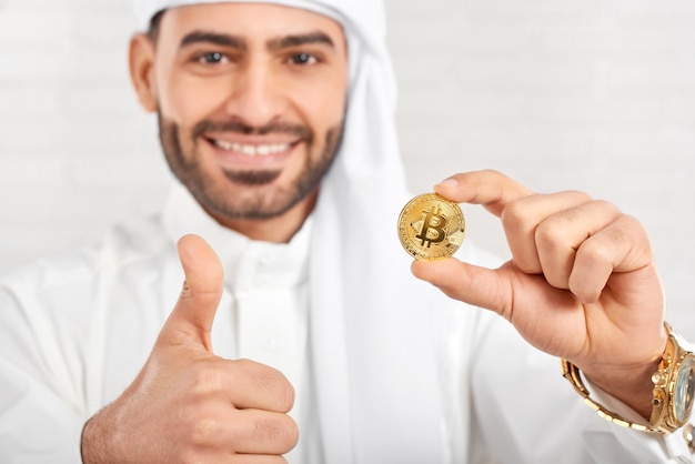 Estúdio close-up de um sorridente empresário árabe mantém bitcoin e parece muito feliz