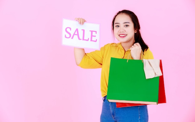 Estúdio de retrato disparou compradora jovem asiática feliz vestindo roupa casual carregando sacolas coloridas, segurando o sinal de papel de banner com desconto de venda, olhando para a câmera sorrindo no fundo rosa.