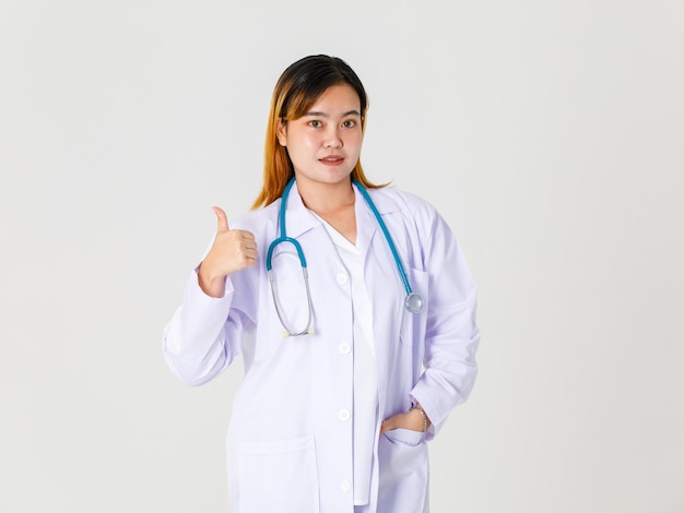 Estúdio de retrato tiro asiático bem sucedido profissional confiante cabelo tingido médico feminino no jaleco pendurado estetoscópio em pé no pescoço, sorrindo, segurando o polegar para cima olhar para a câmera no fundo branco.