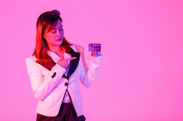 Estúdio de retrato tiro modelo asiático urbano rico rico bem sucedido empresária feminina em tons e terno blazer casual olhar para a câmera segurando o cartão de crédito de compras apontando na mão no fundo rosa.