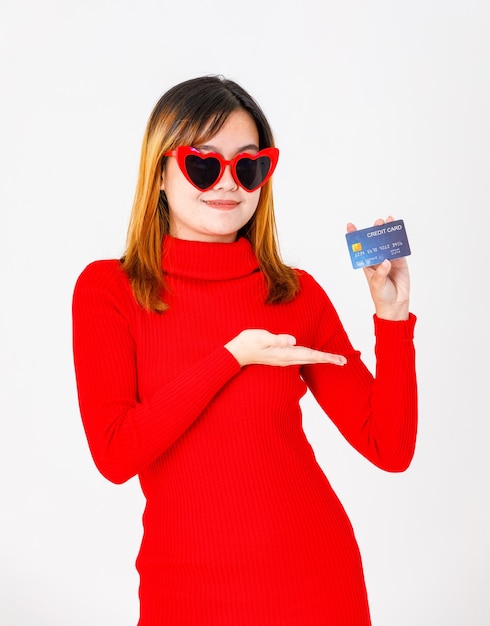 Estudio de retrato disparo Modelo de mujer de negocios asiática de éxito rico rico urbano en vestido rojo y tonos de forma de corazón mira la cámara sosteniendo apuntando con tarjeta de crédito comercial en la mano sobre fondo blanco.