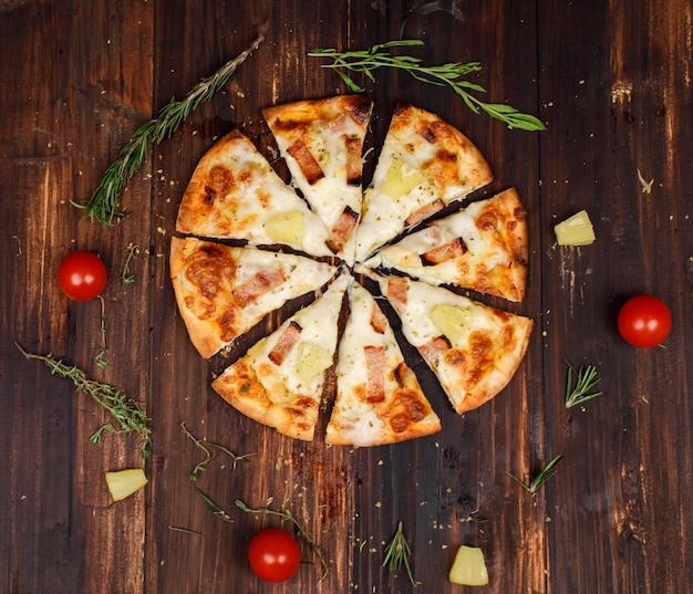 El estudio de la vista superior de primer plano disparó ocho rebanadas al horno sabrosas y deliciosas pizzas de corteza de pan hawaiano tradicional de piña de jamón casero italiano decoradas en una mesa de cocina de madera roja con tomates y hierbas