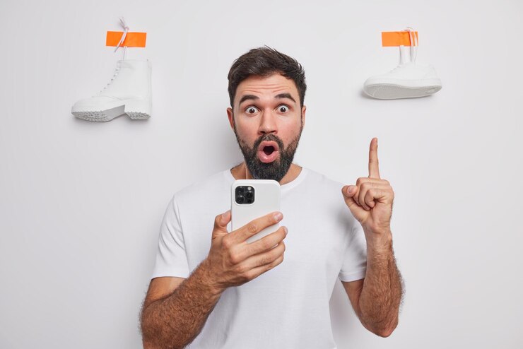 Foto estupefacto joven barbudo mira fijamente los ojos saltones indica que el dedo índice arriba sostiene el teléfono móvil hace compras en línea vestido casualmente posa contra la pared blanca