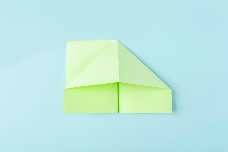 Foto etapa dois de como fazer uma borboleta de papel de origami com papel verde, tesoura em fundo azul