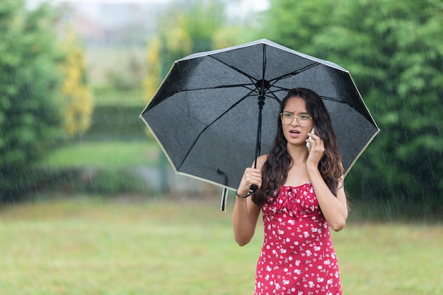 Ethnische Frau mit Regenschirm, die an einem regnerischen Tag im Park telefoniert