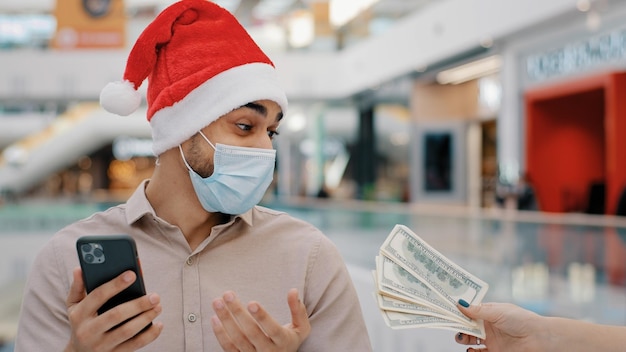 Ethnischer Mann Mann in medizinischer Maske in Weihnachtsweihnachts-Weihnachtsmütze Wetten mit Handy-Casino-Wette