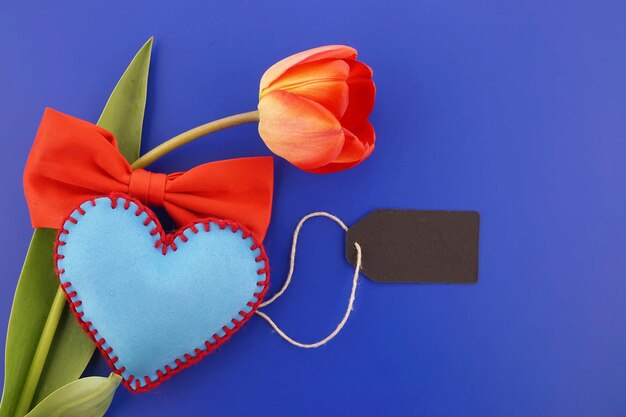 Etiqueta en blanco para un mensaje de San Valentín con corazón de fieltro hecho a mano de tulipán rojo y corbatín sobre azul