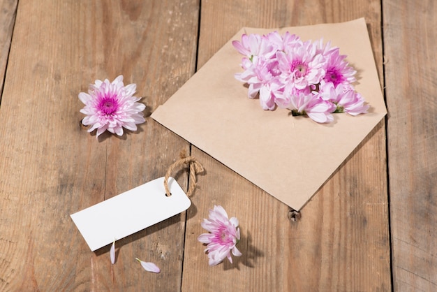 Etiqueta de papel branco em branco com envelope marrom e flores cor de rosa na mesa de madeira.