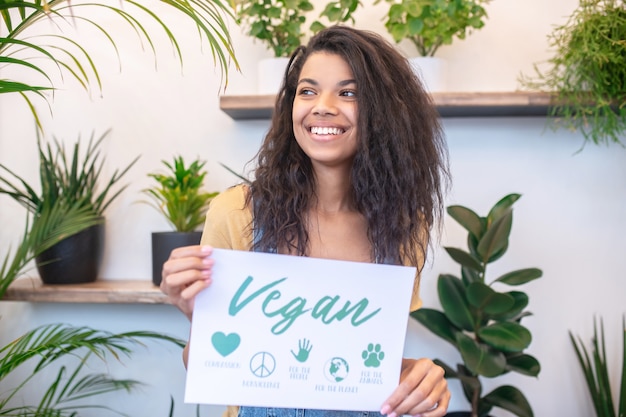 Etiquetado, vegano. Feliz sonriente mujer de cabello oscuro con mirar hacia otro lado sosteniendo un pequeño cartel de marcas veganas