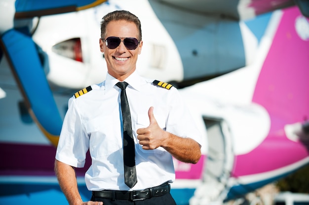 Foto eu amo meu trabalho! piloto confiante do sexo masculino, uniforme, mostrando o polegar para cima e sorrindo