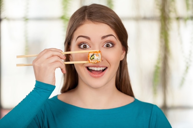 Foto eu amo sushi! mulher jovem e bonita segurando sushi na frente do olho e sorrindo enquanto está sentada no restaurante