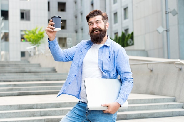 Eu andaria uma milha para tomar um café. Hipster feliz segurar a xícara de café e o laptop. Pausa para o café ao ar livre. Bebida quente. Café para viagem. Nova tecnologia. Vida moderna. Cafe da manha. Experiência rara no paladar.