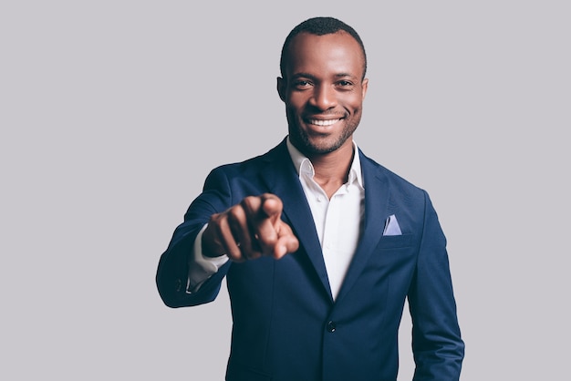 Eu escolho você! Jovem africano bonito em uma jaqueta casual elegante apontando para você e sorrindo em pé