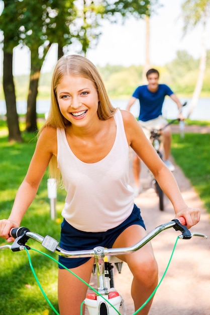 Eu estou ganhando! Mulher jovem e bonita andando de bicicleta e sorrindo enquanto o namorado andando de bicicleta ao fundo