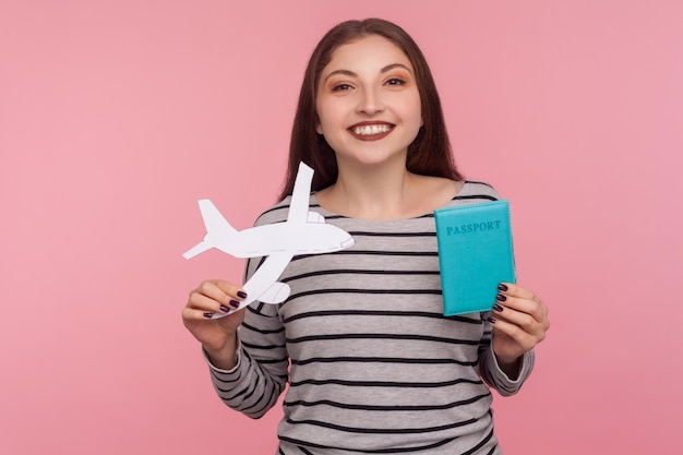 Eu estou indo em viagem. Retrato de mulher jovem e bonita feliz segurando o passaporte e o avião de papel, olhando para a câmera com um sorriso, regozijando-se viajando. tiro de estúdio isolado no fundo rosa