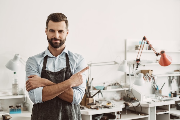 Eu fiz minha escolha e voce Retrato de alegre joalheiro masculino vestindo avental, sorrindo e apontando o dedo para copyspace em pé em seu estúdio de fabricação de joias. O negócio. Oficina de joias