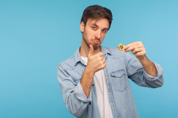 Foto eu gosto de criptomoeda empresário positivo na camisa jeans segurando bitcoin e mostrando os polegares para cima gesto satisfeito com o estúdio de dinheiro eletrônico de moeda btc dourado isolado em fundo azul