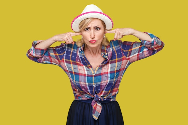 Eu não quero ouvir você. Retrato de mulher madura elegante moderna séria em estilo casual, com chapéu branco em pé, colocando o dedo nas orelhas e olhando. estúdio interno tiro isolado em fundo amarelo.