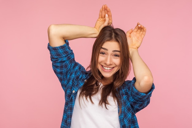 Eu sou coelho. Retrato de uma menina bonita brincalhão incrível na camisa quadriculada, mostrando o gesto de orelhas de coelho, rindo e se divertindo, humor despreocupado infantil. tiro de estúdio interior isolado no fundo rosa