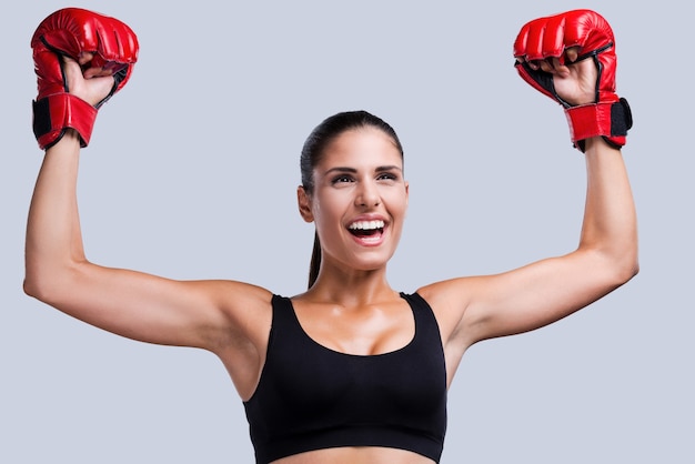 Eu sou um vencedor! Mulher jovem e feliz desportiva com luvas de boxe, mantendo os braços levantados em pé contra um fundo cinza