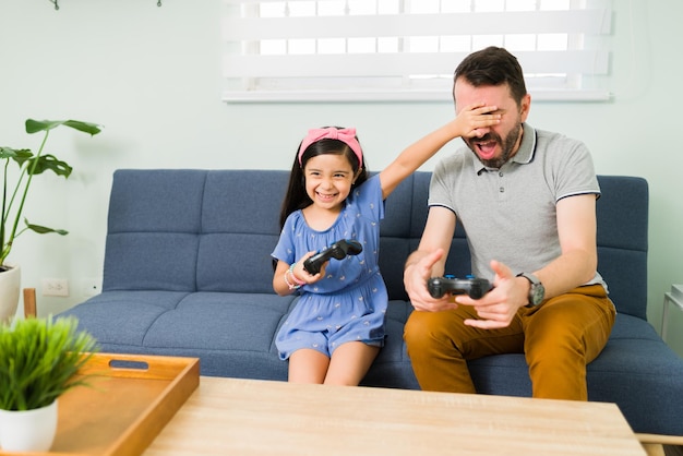 Eu tenho que vencer! Menina bonitinha cobrindo os olhos do pai para impedi-lo de ganhar em um videogame