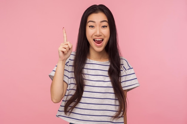 Eureka Retrato de una chica inspirada con el pelo largo y una camiseta a rayas apuntando con el dedo hacia arriba con una idea genial sorprendida por una solución inteligente inventada de repente en un estudio interior aislado en un fondo rosa