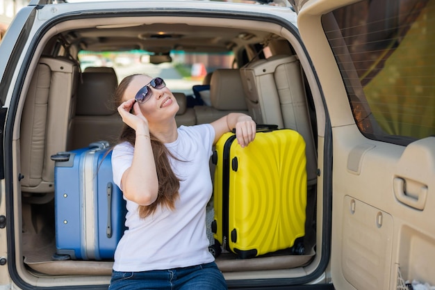 Europäerin mit Sonnenbrille packt ihre Koffer für eine Autofahrt. Schönes glückliches Mädchen sitzt im Kofferraum eines Autos und ist bereit für die Reise. Gelber Koffer mit Dingen für den Sommerurlaub