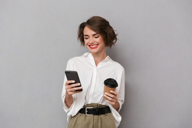 Europäische Frau, die Kaffee zum Mitnehmen hält und Handy verwendet, lokalisiert über graue Wand