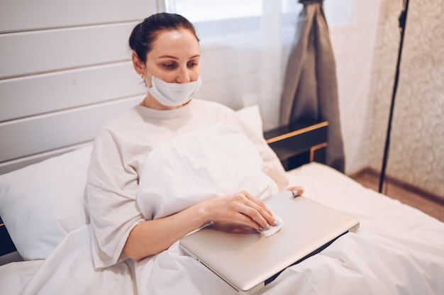 Europäische Frau in der Gesichtsmaske im Schlafzimmer während der Coronavirus-Isolierung nach Hause Quarantäne Reinigung Laptop von Hand Desinfektionsmittel, mit Watte mit Alkohol zu wischen, um eine Kontamination mit Corona-Virus zu vermeiden.