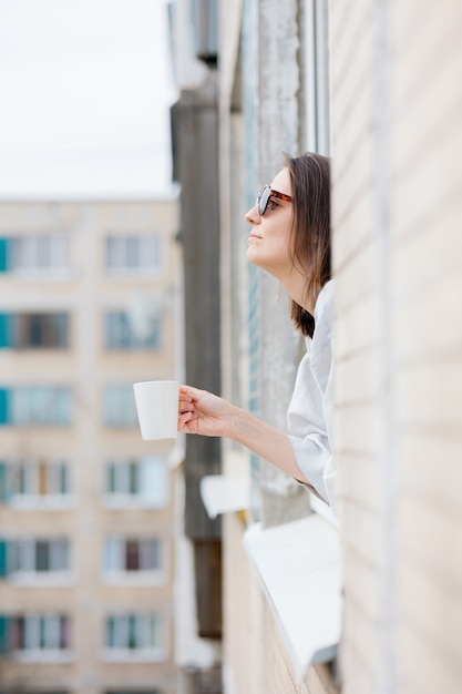 Europäische Frau mit Sonnenbrille und mit einer Tasse Kaffee oder Tee, die aus dem Fenster schaut