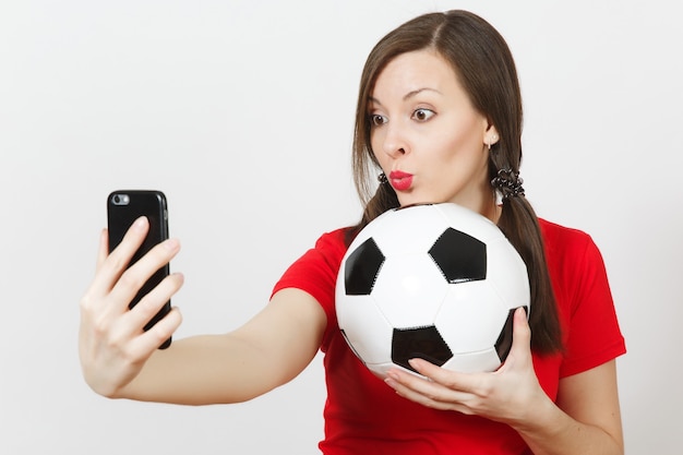 Europäische junge Frau, lustige Pferdeschwänze, Fußballfan oder Spieler in roter Uniform halten Fußball und machen Selfie auf dem Handy einzeln auf weißem Hintergrund. Sport Fußball spielen, gesundes Lifestyle-Konzept.
