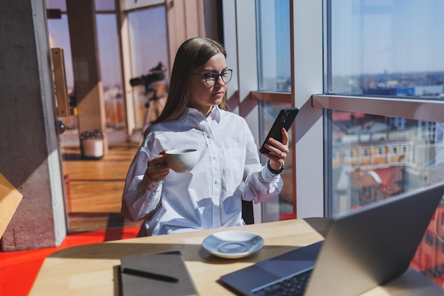 Europäische Managerin sitzt an einem Tisch, während sie an einem Laptop arbeitet und Kaffee in einem Café trinkt Eine junge lächelnde Frau trägt eine Brille und ein weißes Hemd Freiberufliche und Fernarbeit Moderner weiblicher Lebensstil