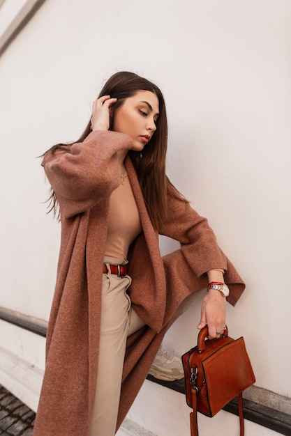 Europäische Mode junge Frau in langem, stylischem Mantel in beige trendiger Hose mit lederbrauner modischer Handtasche in der Nähe von weißem Vintage-Gebäude. Elegantes Mädchenmodell glättet Haare draußen auf der Straße.