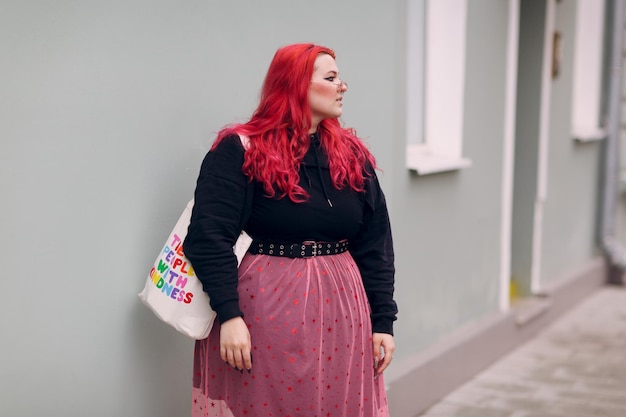 Europäische Plus-Size-Frau. Junges rotrosa behaartes körperpositives Mädchen geht an der Stadtstraße im Freien, die die Mauer umgibt