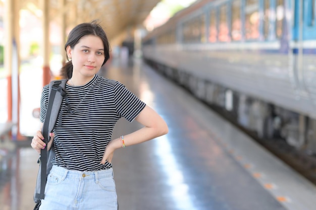 Europäische Teenager-Frau mit Rucksack, die auf dem Bahnhof steht und in die Kamera schaut, gestreiftes T-Shirt und Jeans trägt, Zug rechts geparkt, Zugfahrt.