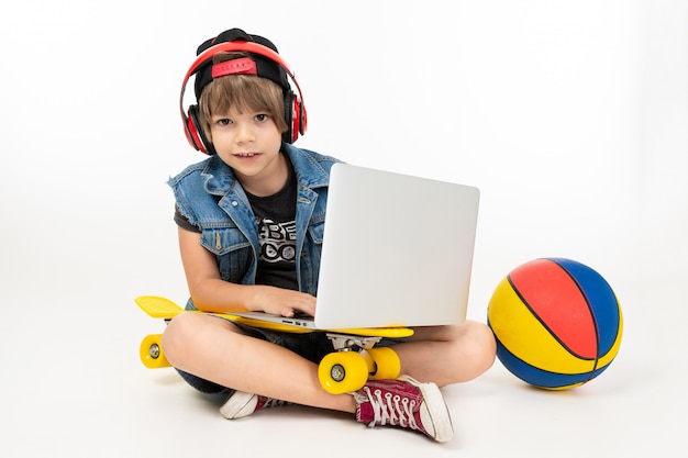 Europäischer Junge, der auf dem Boden mit Laptop, Skateboard und Basketball onwhite sitzt.